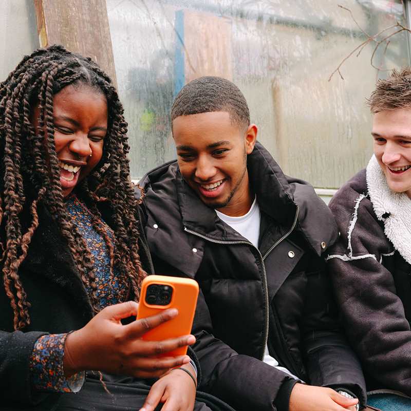 A young Black woman, young Black man and young white man, all sitting on a bench outside, looking at something on a phone and laughing.